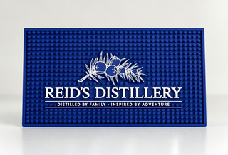 Blue Reid's Distillery bar mat with white juniper artwork and logo, shown against an off-white studio background